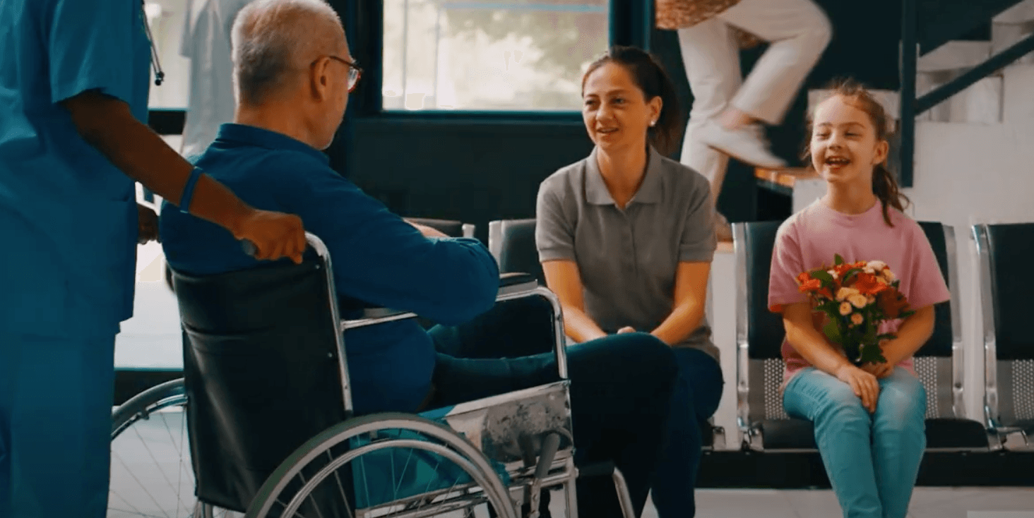 A healthcare worker pushing a man in a wheelchair towards a smiling woman and a girl holding flowers in a waiting area