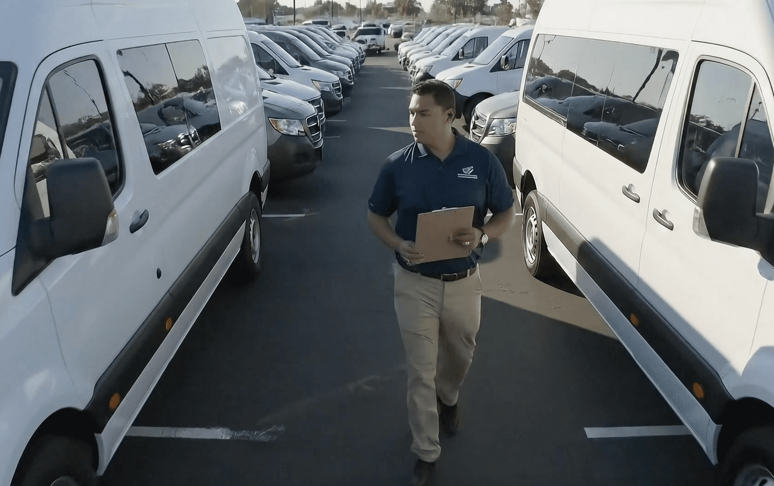 A man with a clipboard walking between rows of white passenger vans in a fleet parking lot